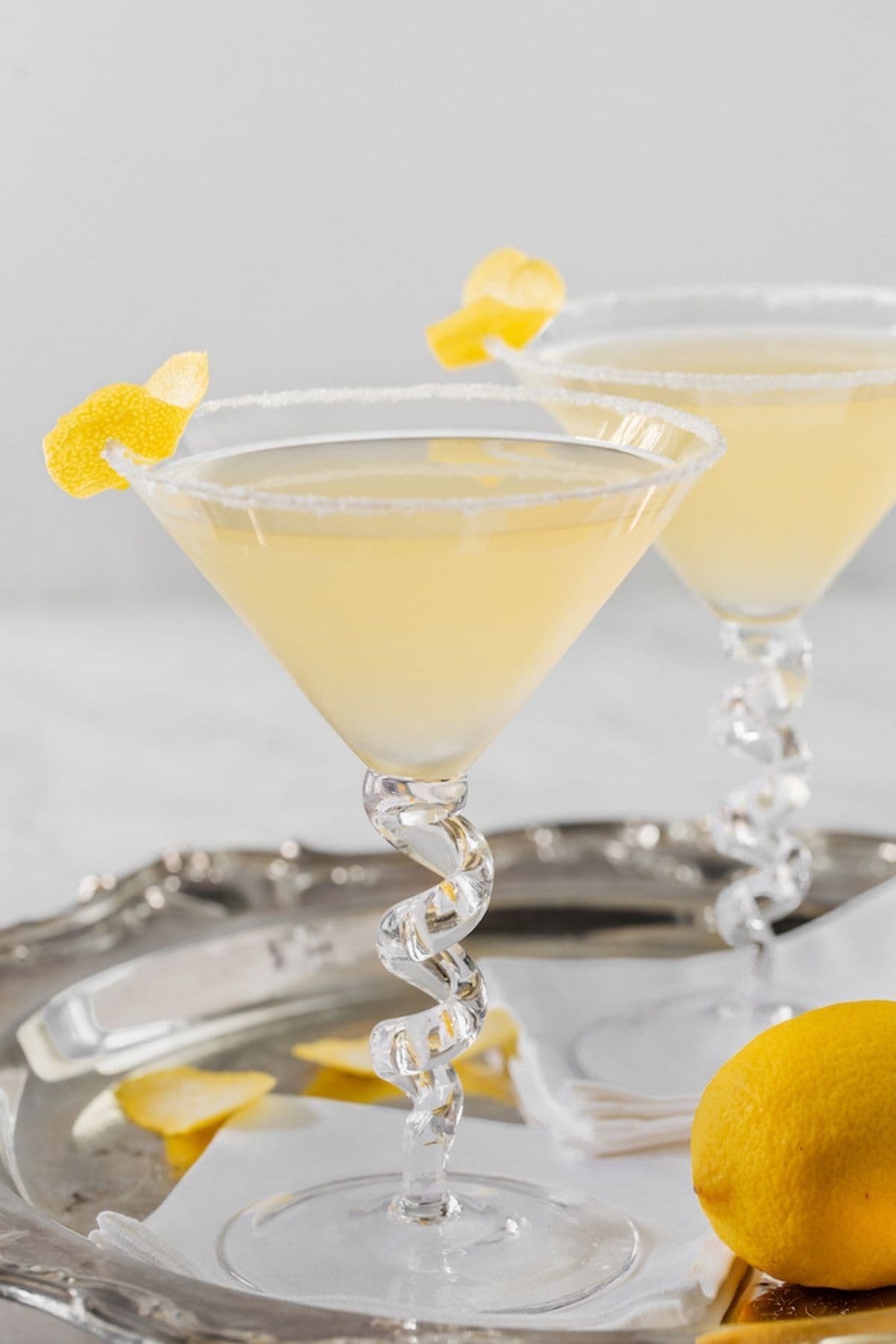 Two martini glasses filled with a lemon drop martini, with sugared rims and lemon twists on the side.