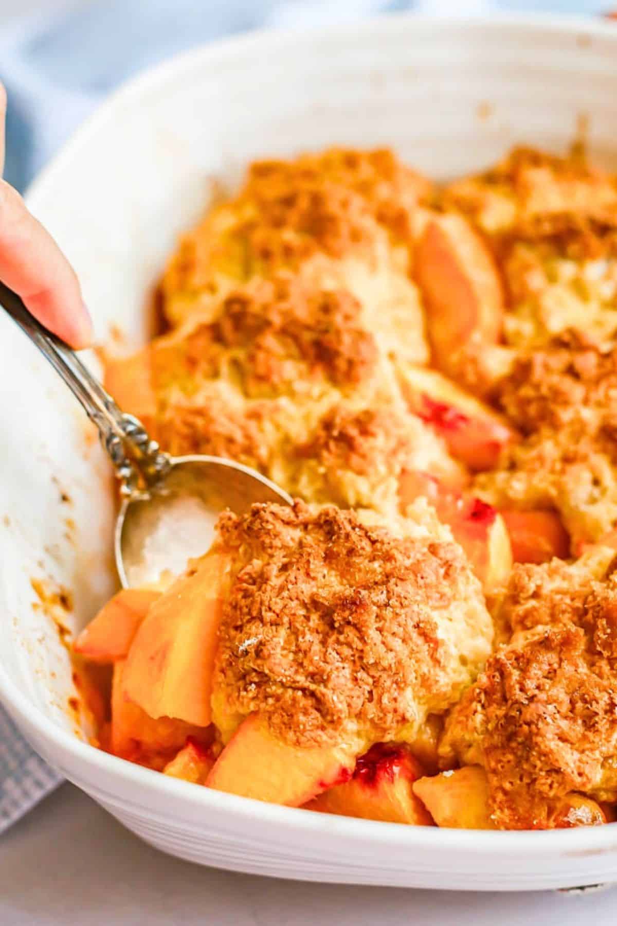 A scoop of peach cobbler being lifted from a white rectangular baking dish.