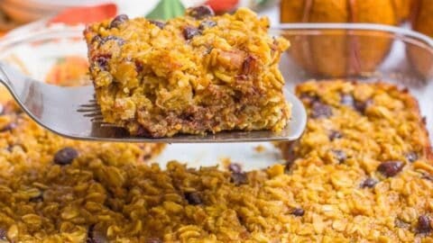 Close up of a spatula lifting up a square piece of pumpkin baked oatmeal with chocolate chips from a pan with plates and decor pumpkins in the background.