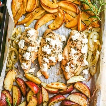 Close up of a sheet pan with chicken breasts topped with goat cheese in the middle and roasted apples to one side and roasted potato wedges to the other side with sprigs of fresh rosemary as garnish.
