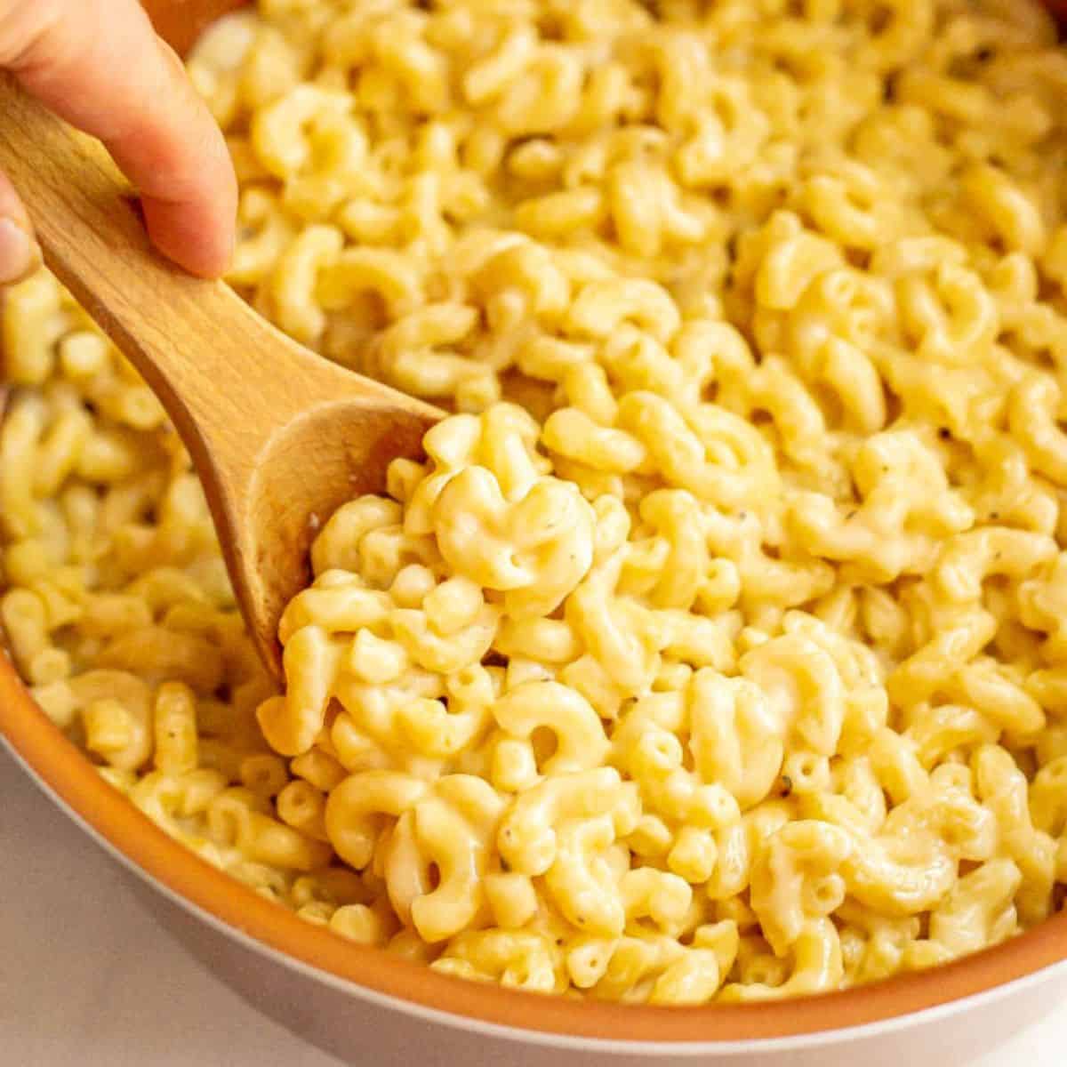 Close up of a hand using a wooden spoon to scoop out some homemade creamy mac and cheese from a large pot.