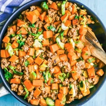 A large skillet filled with a mixture of crumbled turkey sausage, cubed sweet potatoes and sliced Brussels sprouts with a wooden spoon resting in it.