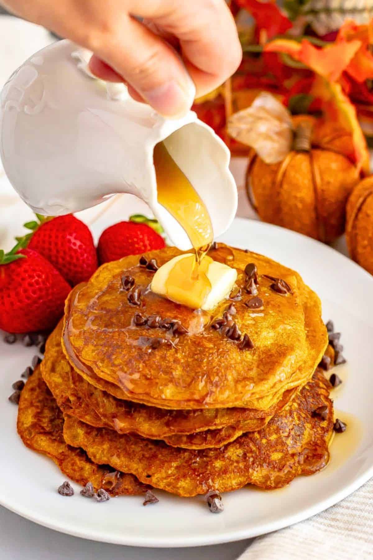 Whole wheat pumpkin pancakes are light, fluffy and full of warm spices and pumpkin flavor. They are naturally sweetened (and can be made vegan) and are perfect for a delicious fall breakfast! #pumpkin #pancakes #breakfast #vegan Maple syrup being poured from a white cruet over a stack of pumpkin pancakes with butter and mini chocolate chips on top.