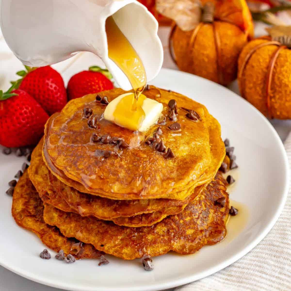 Close up of maple syrup being poured from a white cruet over a stack of pumpkin pancakes with butter and mini chocolate chips on top.