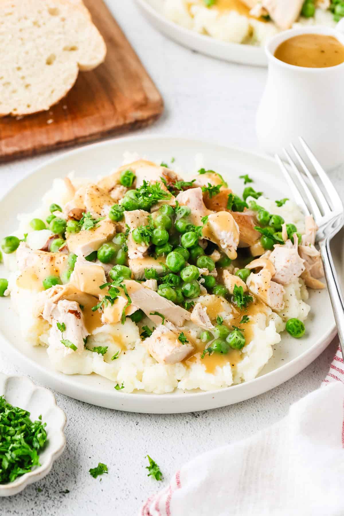 A dinner plate filled with mashed potatoes topped with chicken and peas and drizzled with gravy with a fork to the side.