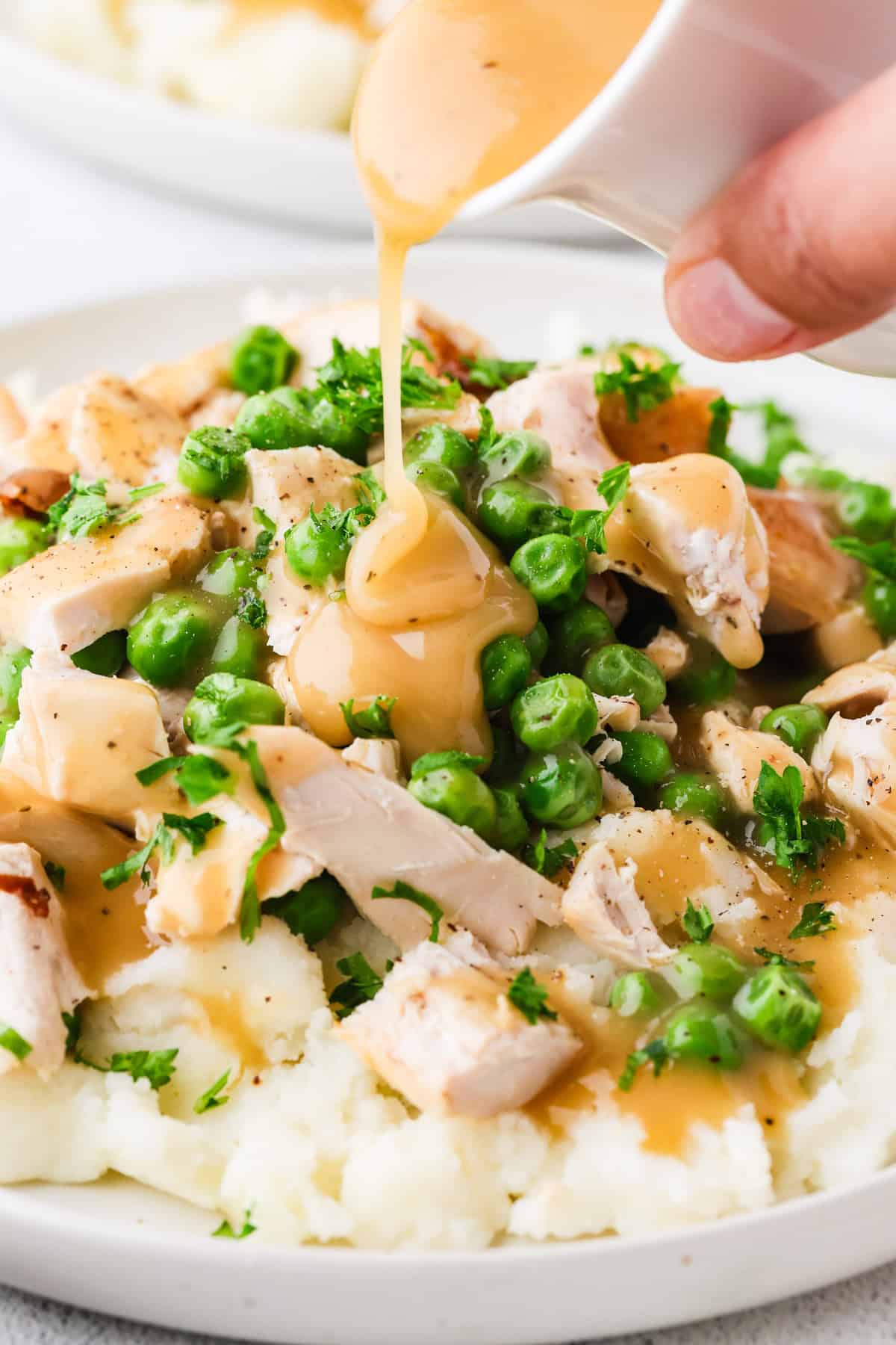 Gravy being poured on top of a mashed potato and chicken bowl with green peas.