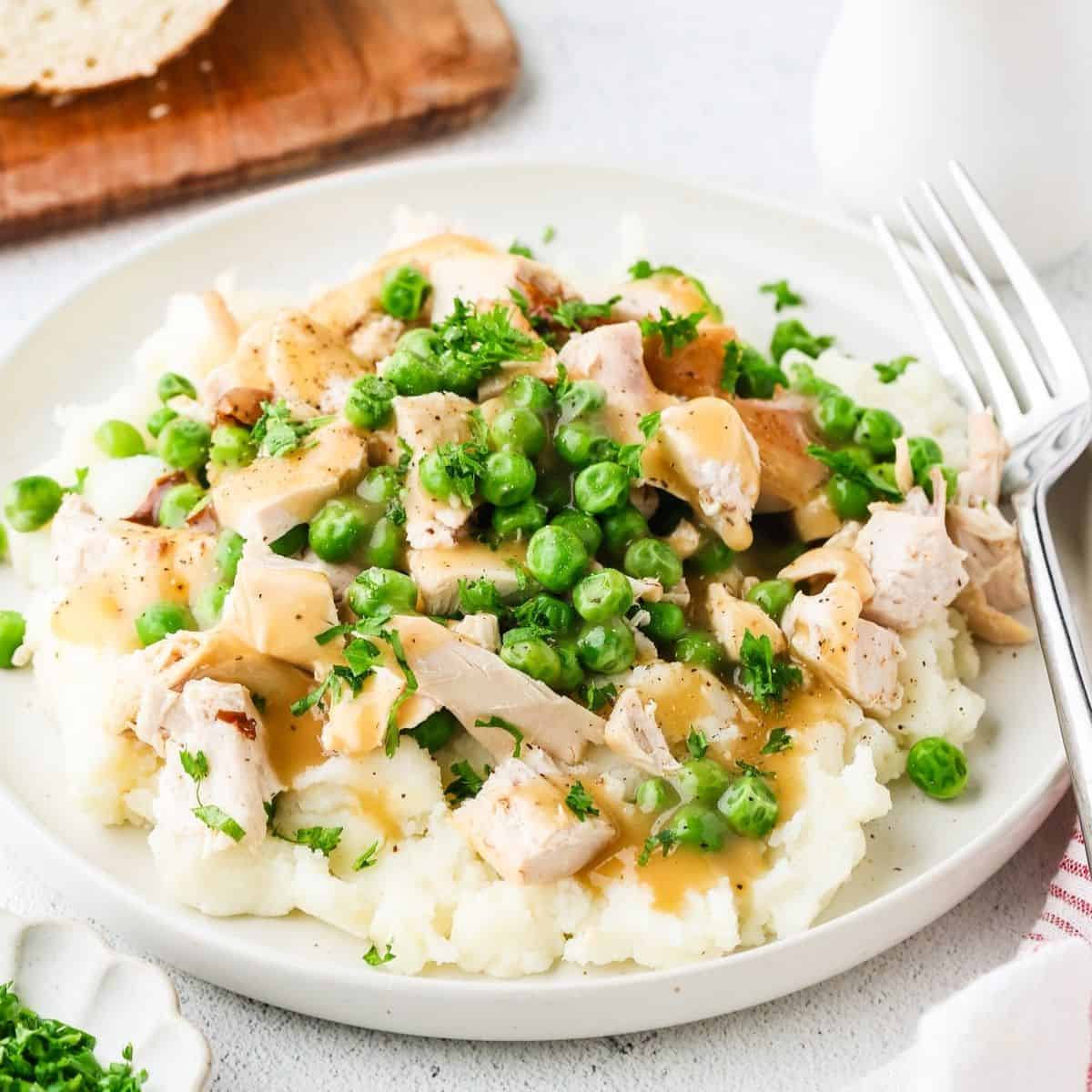 Close up of a dinner plate filled with mashed potatoes topped with chicken and peas and drizzled with gravy with a fork to the side.