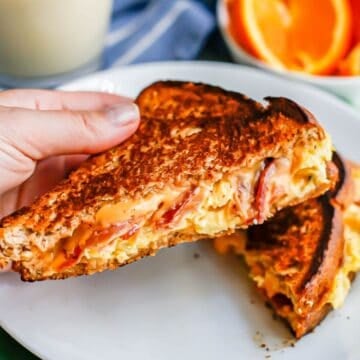 Close up of a hand holding up a half of a breakfast grilled cheese sandwich with scrambled eggs, bacon and cheese.