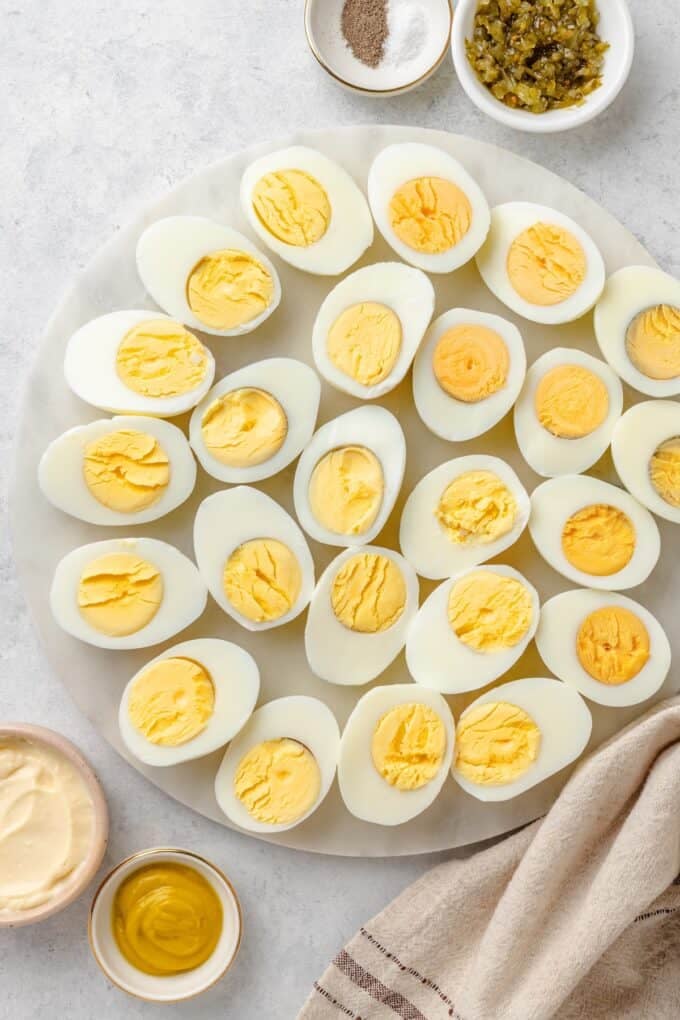 A dozen hard boiled eggs cut in half lengthwise on a round marble platter, with bowls of mayonnaise, mustard, sweet pickle cubes, and salt and pepper nearby.