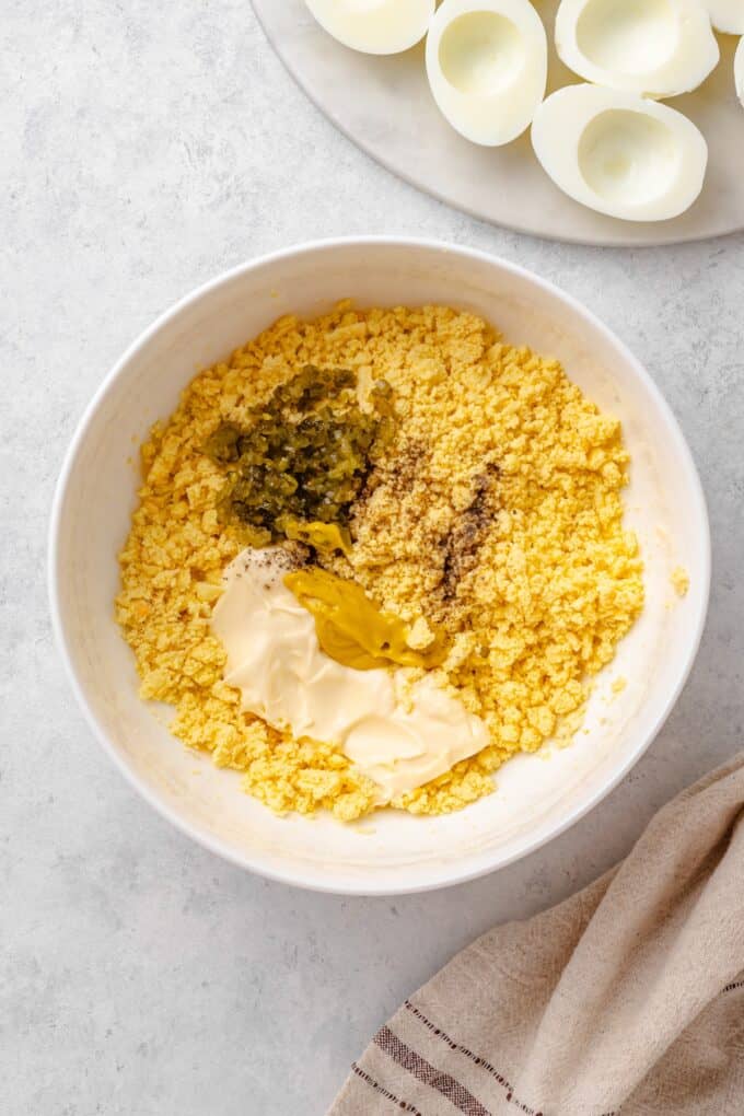 Crumbled egg yolks in a white bowl topped with mayonnaise, yellow mustard, sweet pickle cubes, and salt and pepper, ready to be mixed.