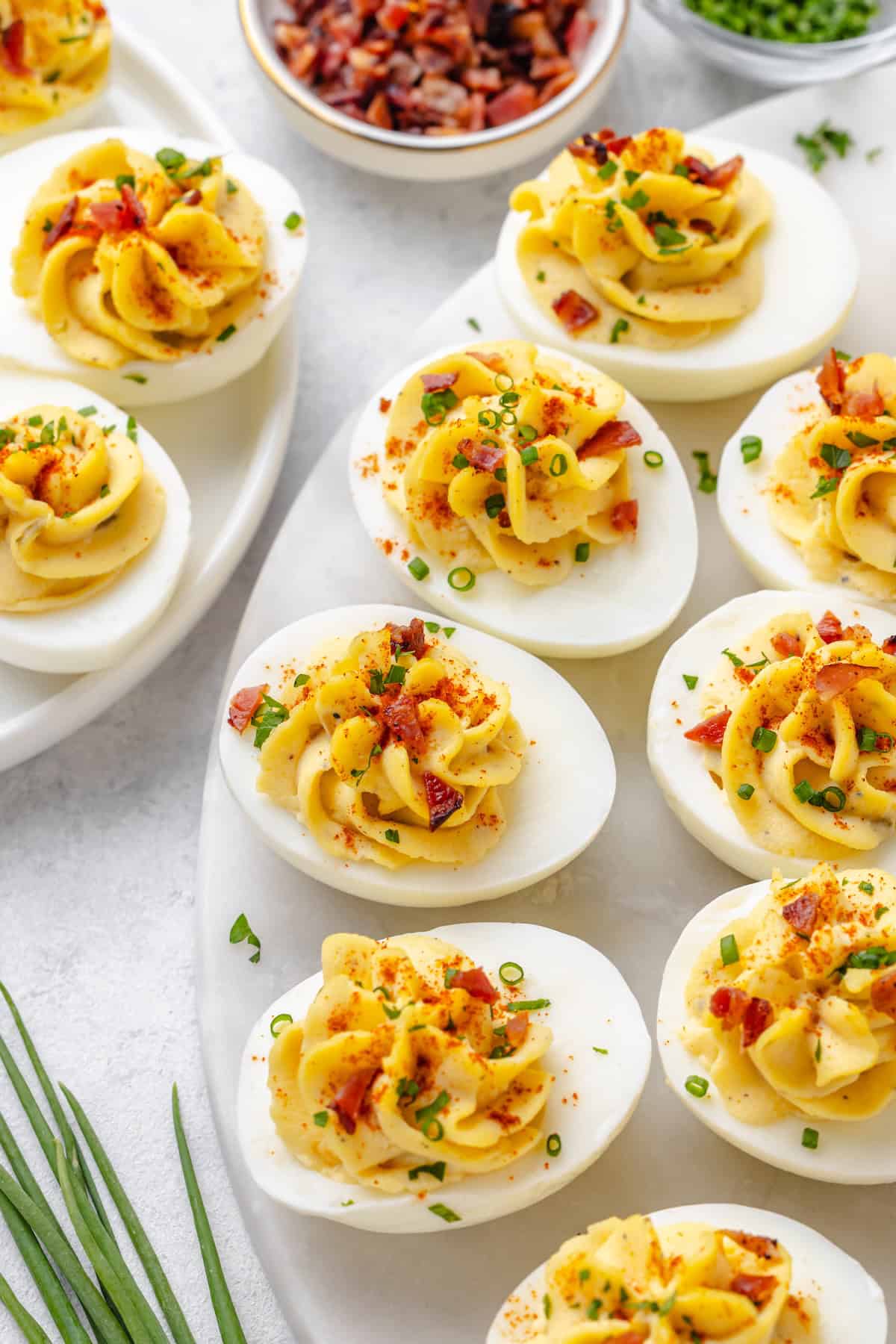 A platter of deviled eggs topped with bacon, chives, and paprika, with small bowls of extra crumbled bacon and chopped chives on the side.