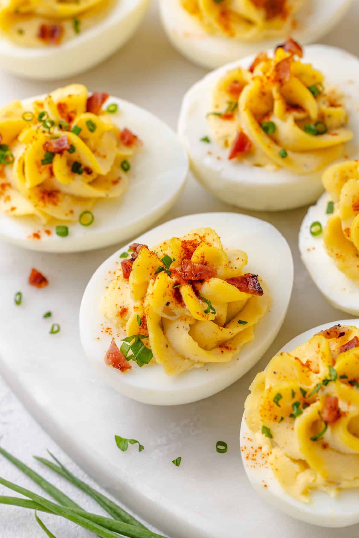 Deviled eggs garnished with bacon, chives, and paprika on a white marble platter, with fresh whole chives to the side.