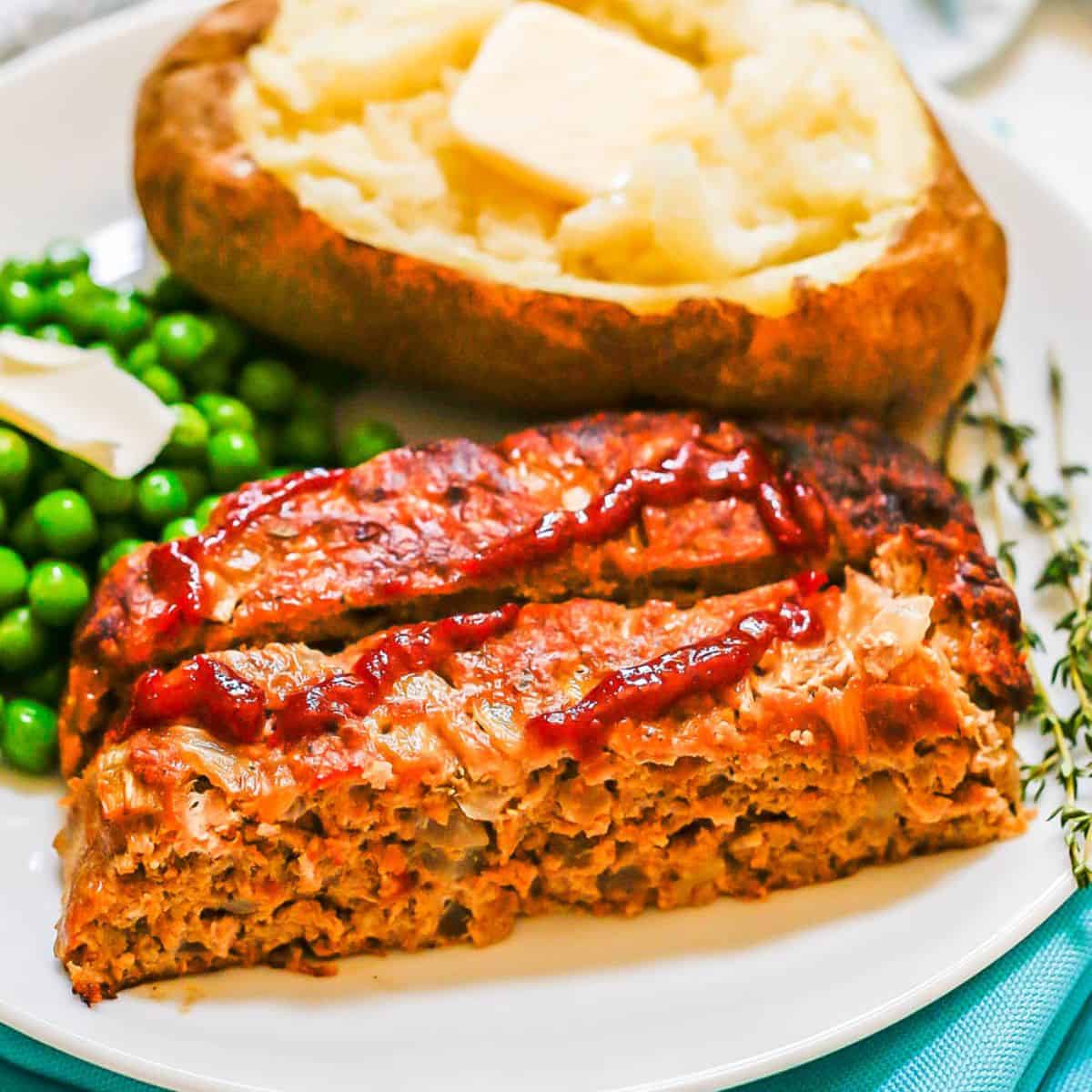 A white round dinner plate with two slices of a turkey meatloaf topped with ketchup with a baked potato and green peas in the background.