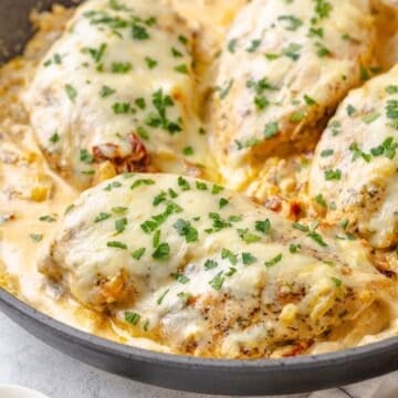 Close up of cheese covered chicken breasts in a creamy sun dried tomato sauce in a large skillet with fresh parsley sprinkled on top.