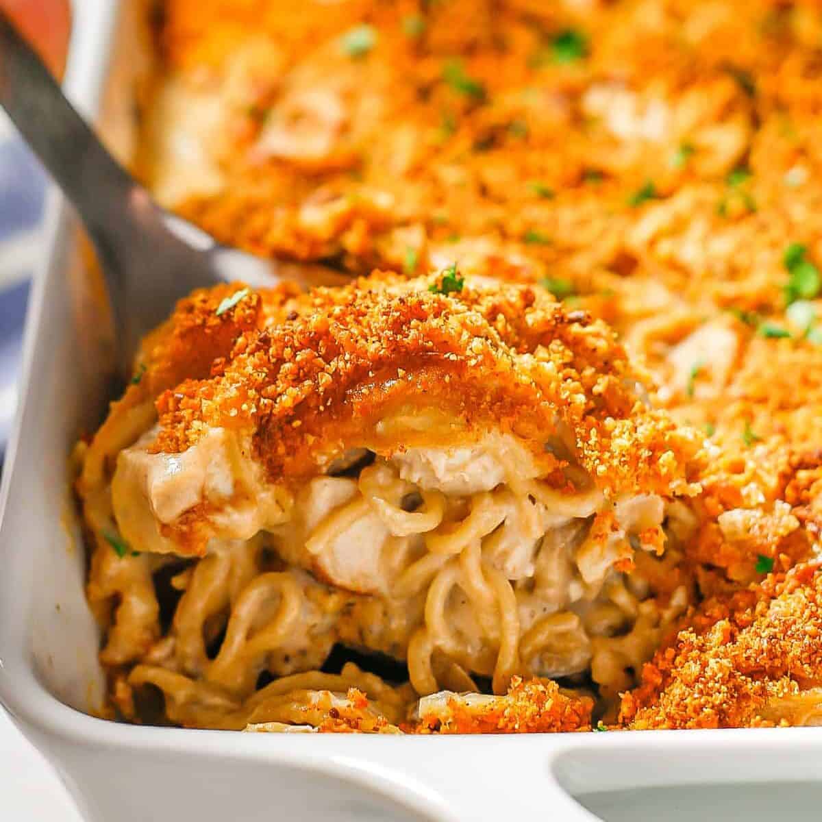 A silver spoon scooping up some creamy turkey tetrazzini with a breadcrumb topping from a white rectangular casserole dish